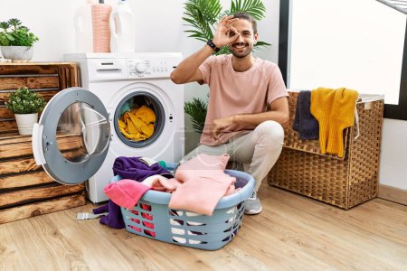 Foto de Young hispanic man putting dirty laundry into washing machine smiling happy doing ok sign with hand on eye looking through fingers - Imagen libre de derechos