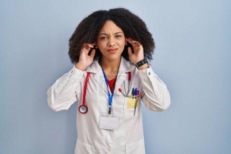 Photo for Young african american woman wearing doctor uniform and stethoscope covering ears with fingers with annoyed expression for the noise of loud music. deaf concept. - Royalty Free Image