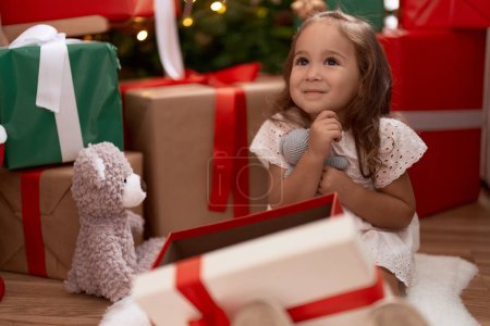 Téléchargez les photos : Adorable fille hispanique étreignant ours en peluche assis sur le sol par l'arbre de Noël à la maison - en image libre de droit
