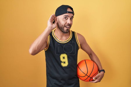 Photo for Middle age bald man holding basketball ball over yellow background smiling with hand over ear listening an hearing to rumor or gossip. deafness concept. - Royalty Free Image