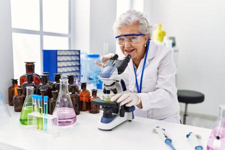 Téléchargez les photos : Femme âgée aux cheveux gris portant un uniforme de scientifique au microscope en laboratoire - en image libre de droit