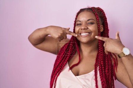 Foto de Mujer afroamericana con el pelo trenzado de pie sobre fondo rosa sonriendo alegre mostrando y señalando con los dedos los dientes y la boca. concepto de salud dental. - Imagen libre de derechos