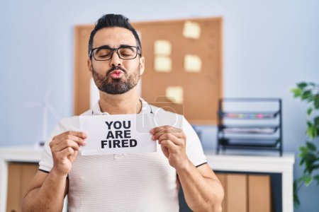 Photo for Young hispanic man with beard holding you are fired banner at the office looking at the camera blowing a kiss being lovely and sexy. love expression. - Royalty Free Image