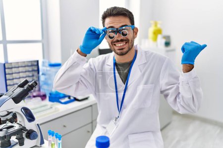 Photo for Young hispanic man working at scientist laboratory wearing magnifying glasses pointing thumb up to the side smiling happy with open mouth - Royalty Free Image