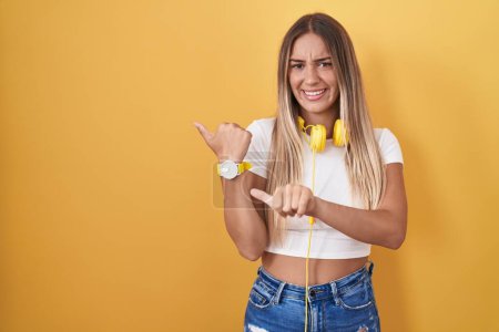 Foto de Mujer rubia joven de pie sobre fondo amarillo con auriculares apuntando a la espalda con la mano y los pulgares hacia arriba, sonriendo confiado - Imagen libre de derechos