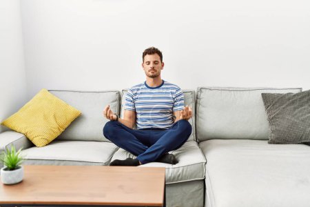 Photo for Young hispanic man doing yoga exercise sitting on the sofa at home - Royalty Free Image