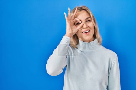 Photo for Young caucasian woman standing over blue background doing ok gesture with hand smiling, eye looking through fingers with happy face. - Royalty Free Image