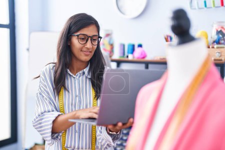 Téléchargez les photos : Jeune belle femme hispanique tailleur souriant confiant en utilisant un ordinateur portable à l'usine de vêtements - en image libre de droit