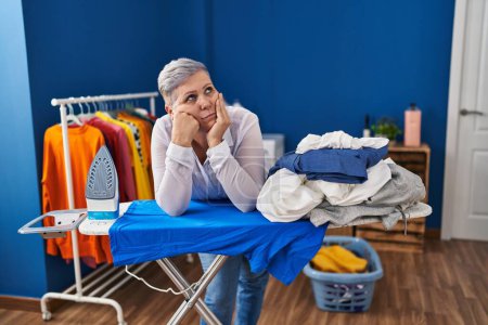 Foto de Mujer de mediana edad aburrida apoyada en la tabla de planchar en la lavandería - Imagen libre de derechos