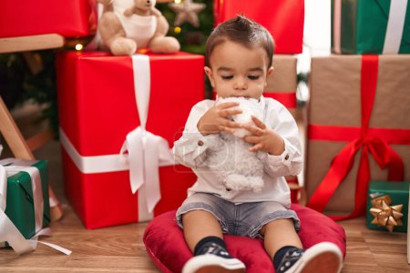 Photo for Adorable hispanic toddler hugging teddy bear sitting on floor by christmas gifts at home - Royalty Free Image