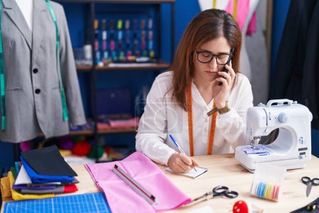 Téléchargez les photos : Young woman tailor talking on smartphone writing on notebook at sewing studio - en image libre de droit