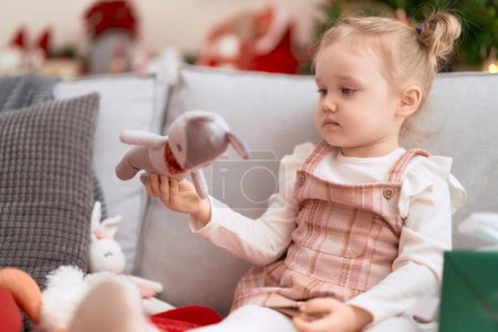 Photo for Adorable caucasian girl holding toy sitting on sofa by christmas tree at home - Royalty Free Image