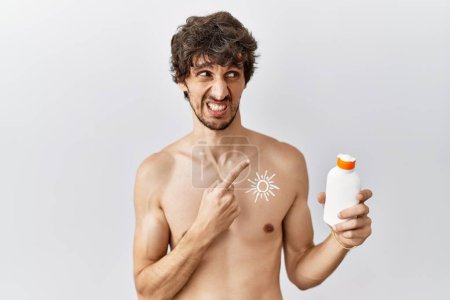 Téléchargez les photos : Jeune homme hispanique debout torse nu tenant crème solaire lotion pointant de côté inquiet et nerveux avec index, expression inquiète et surprise - en image libre de droit
