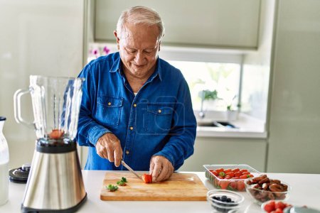 Foto de Hombre mayor sonriendo confiado cortando fresa en la cocina - Imagen libre de derechos