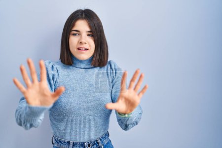 Photo for Young hispanic woman standing over blue background afraid and terrified with fear expression stop gesture with hands, shouting in shock. panic concept. - Royalty Free Image
