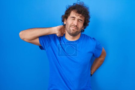 Téléchargez les photos : Jeune homme hispanique debout sur fond bleu souffrant d'une blessure au cou, toucher le cou avec la main, douleur musculaire - en image libre de droit