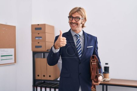 Photo for Caucasian man with mustache working at the office wearing glasses smiling happy and positive, thumb up doing excellent and approval sign - Royalty Free Image