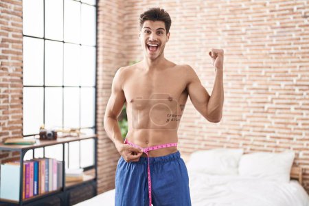Photo for Young hispanic man using tape measure measuring waist screaming proud, celebrating victory and success very excited with raised arm - Royalty Free Image