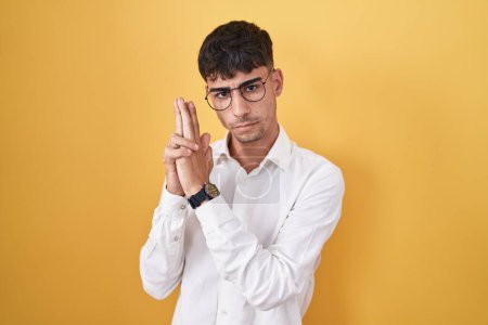 Photo for Young hispanic man standing over yellow background holding symbolic gun with hand gesture, playing killing shooting weapons, angry face - Royalty Free Image