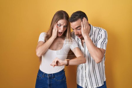 Téléchargez les photos : Jeune couple debout sur fond jaune regardant le temps de la montre inquiet, peur de se faire tard - en image libre de droit