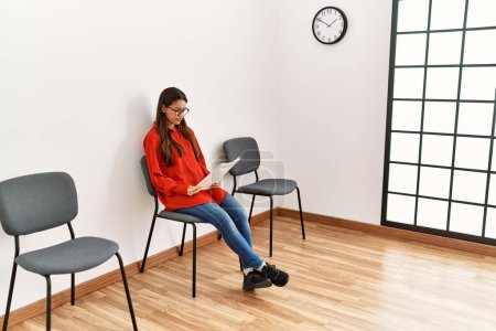 Téléchargez les photos : Jeune femme latine lisant un document assis sur une chaise dans une salle d'attente - en image libre de droit