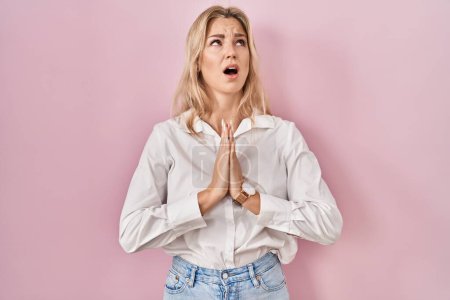 Téléchargez les photos : Jeune femme caucasienne portant une chemise blanche décontractée sur fond rose mendiant et priant avec les mains ainsi que l'expression d'espoir sur le visage très émotionnel et inquiet. mendier. - en image libre de droit