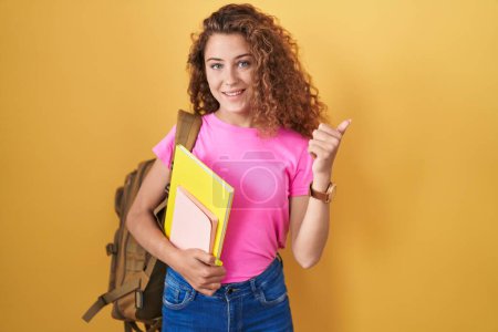 Foto de Young caucasian woman wearing student backpack and holding books doing happy thumbs up gesture with hand. approving expression looking at the camera showing success. - Imagen libre de derechos