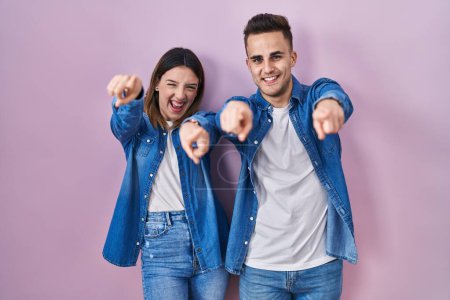 Foto de Joven pareja hispana de pie sobre fondo rosa señalándote a ti y a la cámara con los dedos, sonriendo positiva y alegre - Imagen libre de derechos