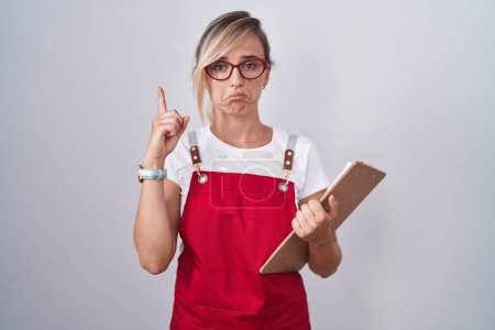 Téléchargez les photos : Young blonde woman wearing waiter uniform holding clipboard pointing up looking sad and upset, indicating direction with fingers, unhappy and depressed. - en image libre de droit