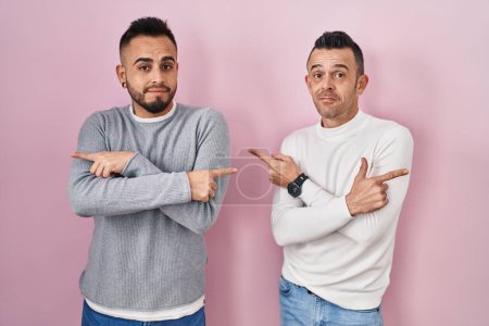 Téléchargez les photos : Couple homosexuel debout sur fond rose pointant des deux côtés avec les doigts, direction différente en désaccord - en image libre de droit
