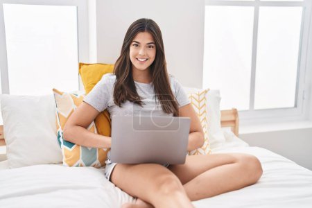 Photo for Hispanic woman using computer laptop on the bed looking positive and happy standing and smiling with a confident smile showing teeth - Royalty Free Image