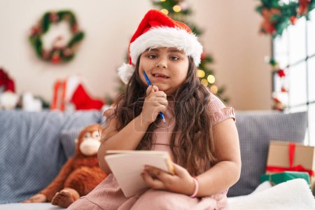 Photo for Plus size hispanic girl thinking to write sitting on sofa by christmas tree at home - Royalty Free Image