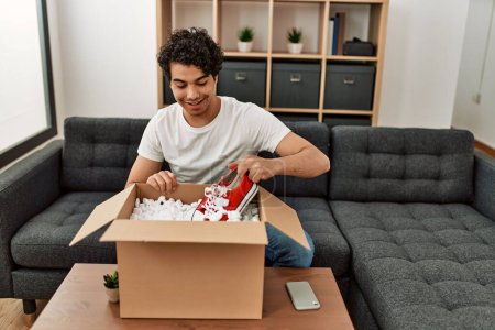 Photo for Young hispanic man unboxing sneakers of cardboard box sitting on the sofa at home. - Royalty Free Image