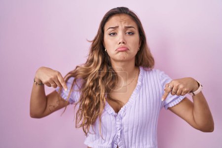 Téléchargez les photos : Young hispanic woman standing over pink background pointing down looking sad and upset, indicating direction with fingers, unhappy and depressed. - en image libre de droit