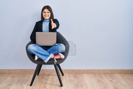 Téléchargez les photos : Jeune femme hispanique assise sur une chaise en utilisant ordinateur portable souriant amical offrant poignée de main comme salutation et accueil. entreprise prospère. - en image libre de droit