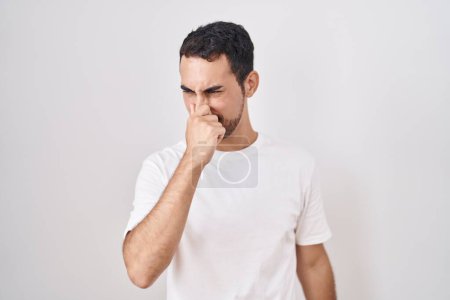 Handsome hispanic man standing over white background smelling something stinky and disgusting, intolerable smell, holding breath with fingers on nose. bad smell 