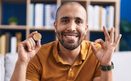 Téléchargez les photos : Hispanic man with beard holding virtual currency bitcoin doing ok sign with fingers, smiling friendly gesturing excellent symbol - en image libre de droit