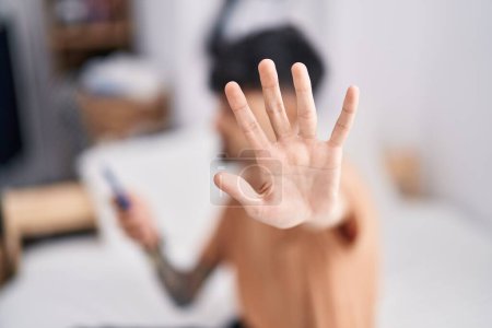 Photo for Young hispanic man using smartphone doing stop gesture with hand at bedroom - Royalty Free Image