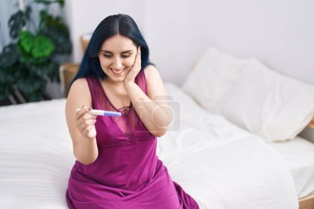 Foto de Joven mujer caucásica sonriendo confiado sosteniendo prueba de embarazo en el dormitorio - Imagen libre de derechos