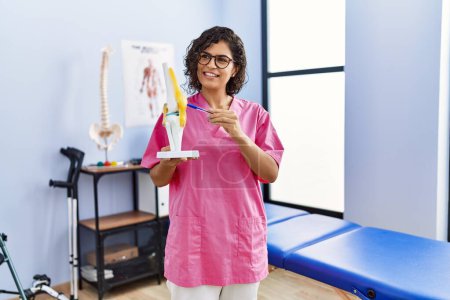 Téléchargez les photos : Jeune femme latine portant un uniforme de physiothérapeute tenant le modèle anatomique du genou à la clinique - en image libre de droit