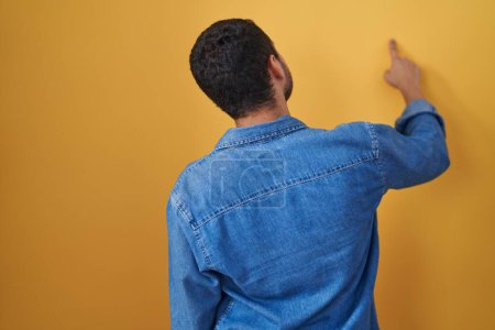 Photo for Hispanic man standing over yellow background posing backwards pointing ahead with finger hand - Royalty Free Image