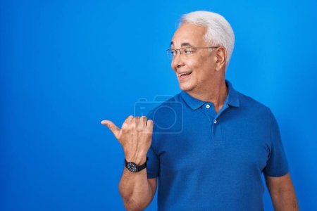 Téléchargez les photos : Homme d'âge moyen avec les cheveux gris debout sur fond bleu souriant avec le visage heureux regardant et pointant vers le côté avec le pouce vers le haut. - en image libre de droit