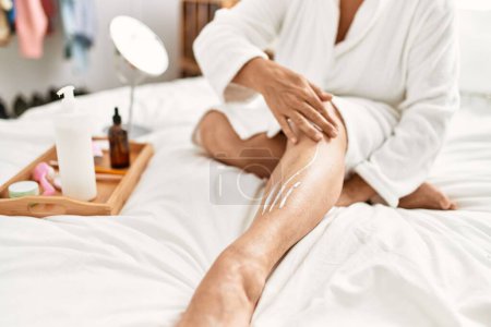 Foto de Woman caring skin legs using hydration cream sitting on the bed at bedroom. - Imagen libre de derechos