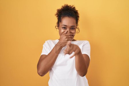Photo for Young hispanic woman with curly hair standing over yellow background laughing at you, pointing finger to the camera with hand over mouth, shame expression - Royalty Free Image