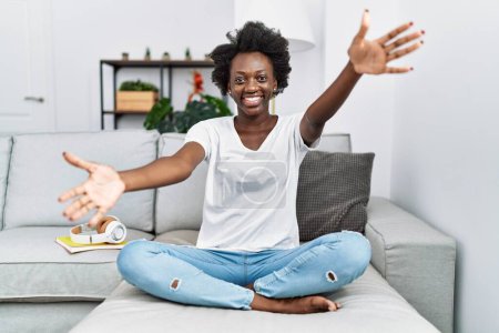 Photo for African young woman sitting on the sofa at home looking at the camera smiling with open arms for hug. cheerful expression embracing happiness. - Royalty Free Image