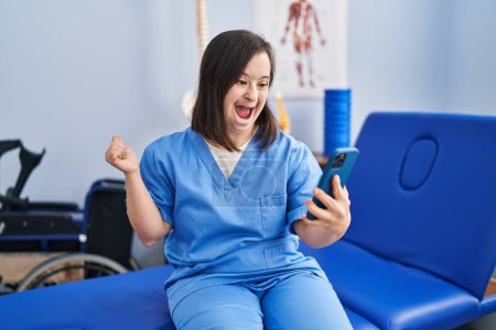Photo for Hispanic girl with down syndrome working at physiotherapy using smartphone screaming proud, celebrating victory and success very excited with raised arm - Royalty Free Image