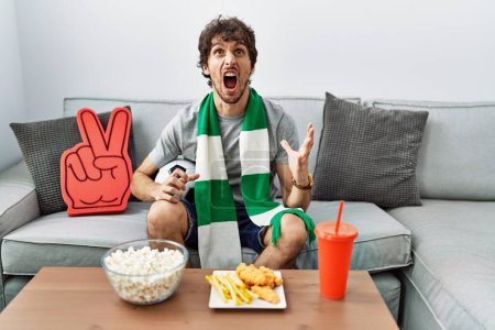 Photo for Young hispanic man football hooligan cheering game at home crazy and mad shouting and yelling with aggressive expression and arms raised. frustration concept. - Royalty Free Image