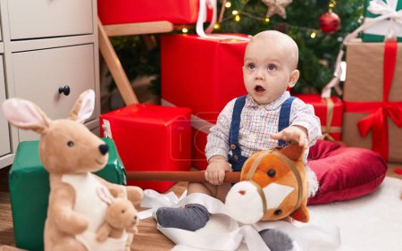 Foto de Adorable bebé caucásico sosteniendo juguete de caballo sentado en el suelo por el árbol de Navidad en casa - Imagen libre de derechos