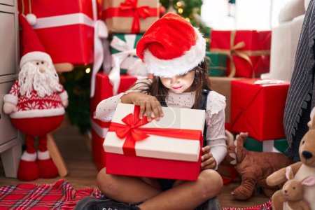 Photo for Adorable hispanic girl unpacking gift sitting on floor by christmas tree at home - Royalty Free Image