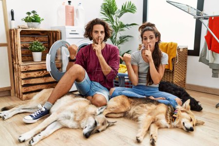 Photo for Young hispanic couple doing laundry with dogs asking to be quiet with finger on lips pointing with hand to the side. silence and secret concept. - Royalty Free Image